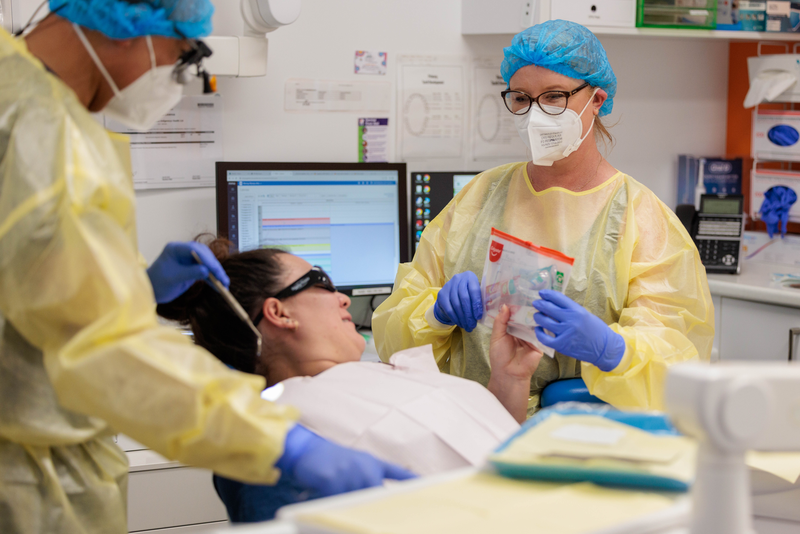 A dentist and dental assist with a patient undergoing a dental exam, wearing a yellow disposable gown, mask, eye glasses and gloves.
