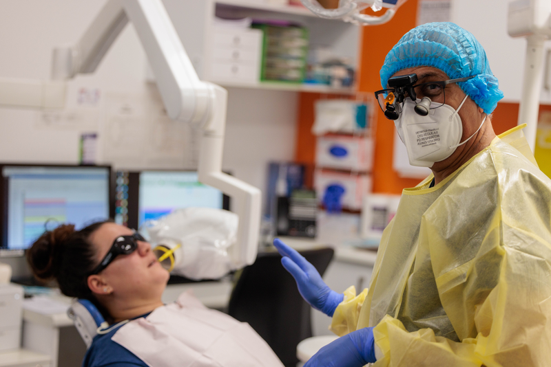 A dentist with a patient undergoing a dental and oral health exam, the dentist is looking at the camera and is wearing a yellow disposable gown, mask, eye glasses and gloves.