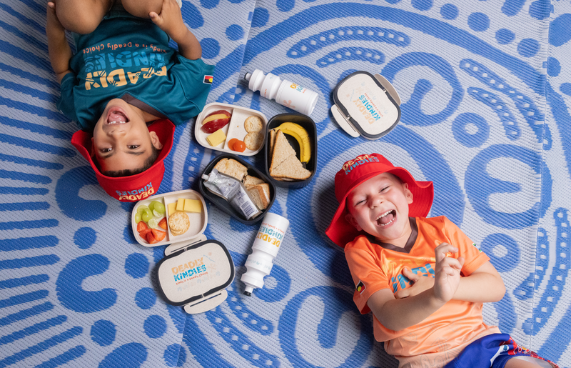 A playgroup with two young Indigenous children lying on a mat with Indigenous artwork designs, there is lunchboxes filled with healthy foods and labels saying Deadly Kindies for Indigenous early childhood education.