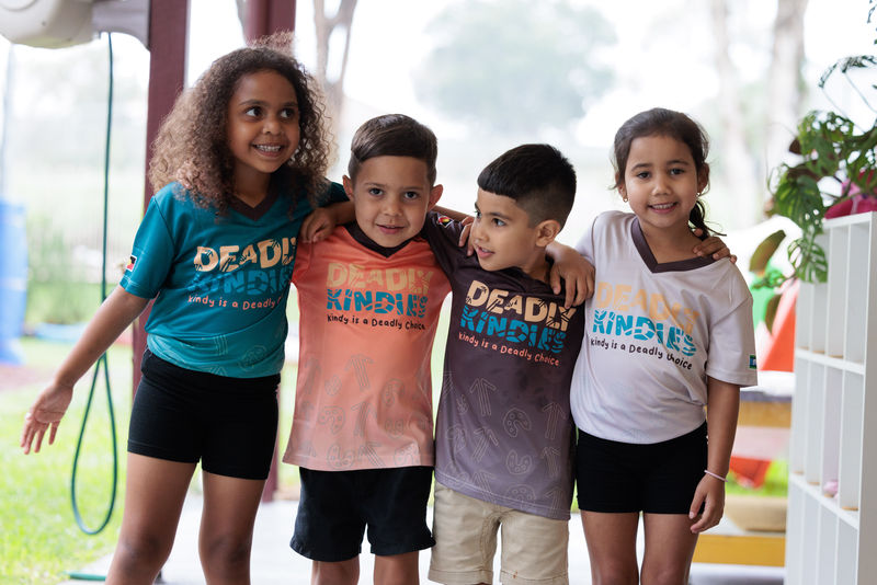 Four young children stand arm in arm, smiling at the camera. They wear colourful shirts with DEADLY KINDIES printed on them, celebrating Indigenous early childhood education in Goodna. The blurred background shows trees and playground equipment.