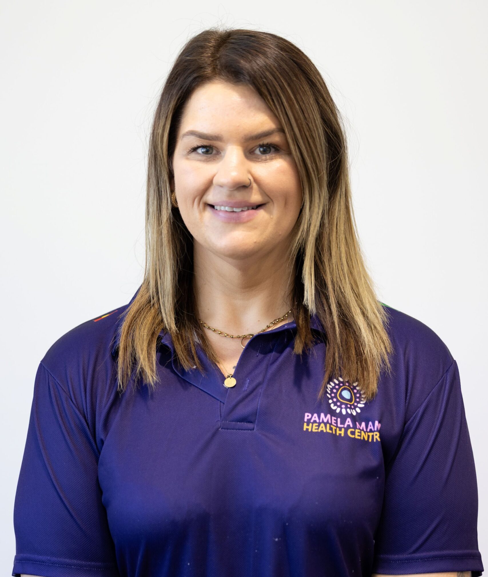 A woman with straight, shoulder-length brown hair, wearing a dark blue polo shirt with "Pamela Mam Health Centre" logo, stands in front of a plain white background and smiles at the camera.