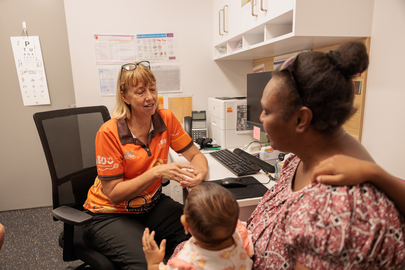 A healthcare worker discusses child health for Mob in Goodna with a woman holding a baby during a clinic consultation, smiling warmly and gesturing as the woman and baby listen attentively.