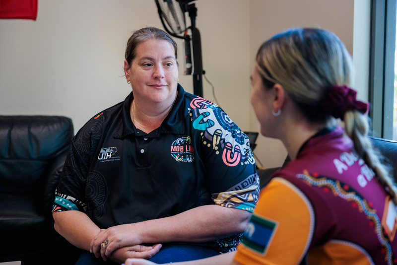 Two women sit facing each other in conversation. One wears a black Mob Link shirt with colourful Indigenous patterns, reflecting care coordination for Mob in Goodna, while the other wears a maroon and yellow sports jersey. They are indoors, sitting near a window.