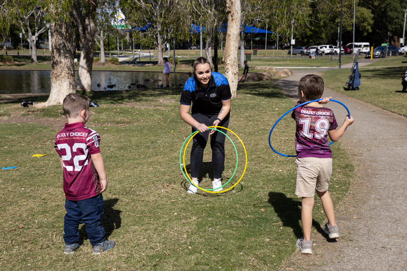 A disability worker smiles while holding two hula hoops, interacting with two young boys wearing maroon sports jerseys in a sunny park, highlighting Disability and NDIS support for Mob in Goodna amid trees, grass, and a pond in the background.