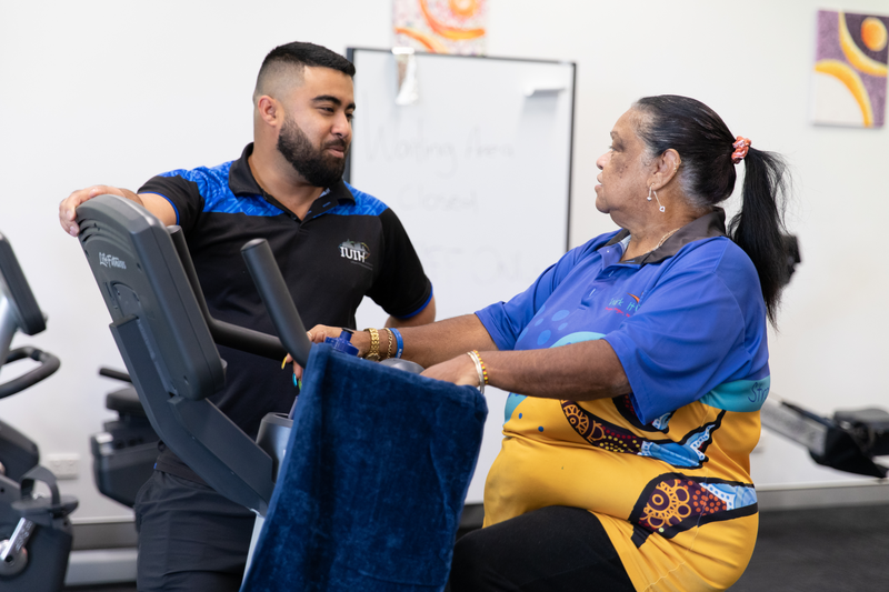 A exercise physiologist with an Indigenous client who is wearing the Work It Out program uniform. She is on an exercise bike at a local gym.