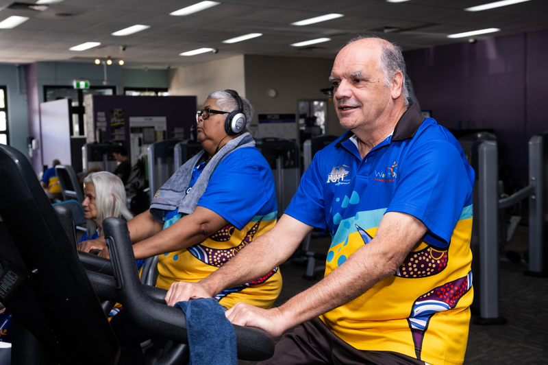 Two older adults wearing colourful shirts exercise on cardio machines at a gym, showcasing the benefits of exercise rehab for Mob in Goodna. The man in front smiles, while the woman beside him wears headphones and a towel over her shoulder.