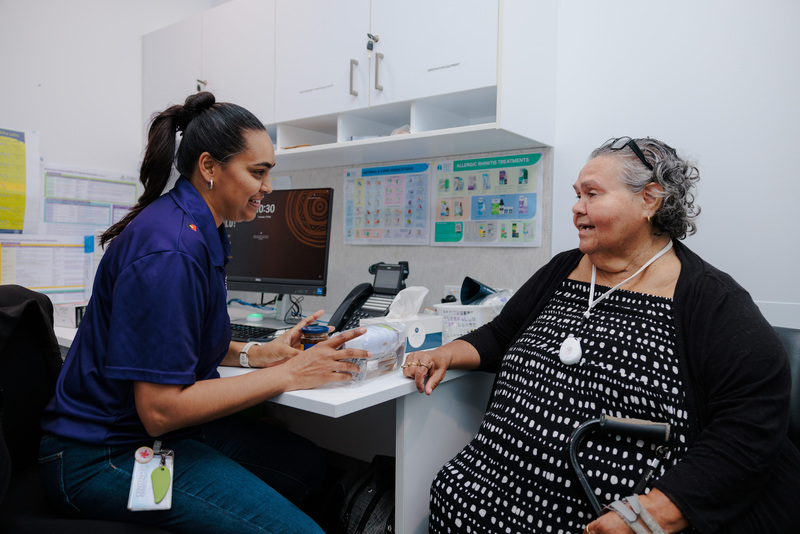 A healthcare worker sits at a desk, talking with an older woman holding a cane. The healthcare worker gestures while explaining something, and both appear engaged in conversation in a medical office setting about Occupational Therapy.