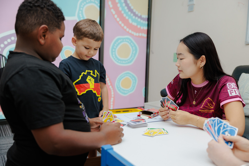 A speech pathologist sits with two children, they are playing UNO (card games) at a table together with Aboriginal artwork in the background.
