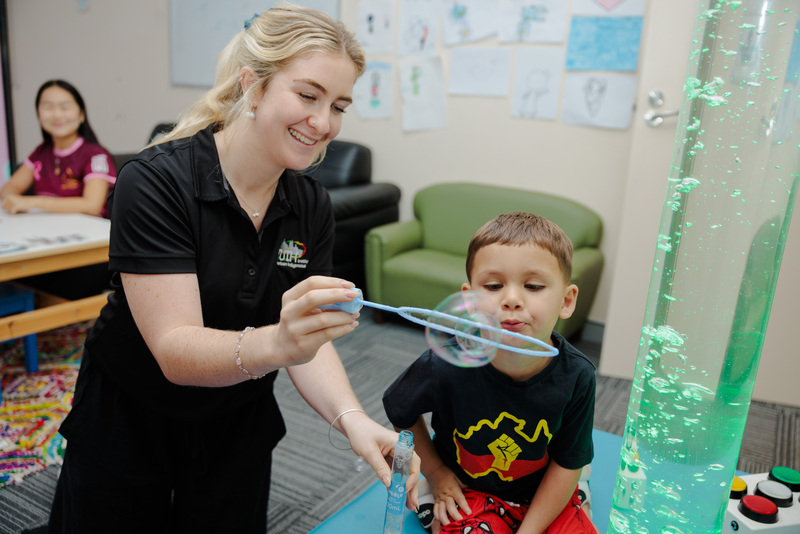 A speech pathologist and a child blow bubbles together, another staff member sits in the background smiling as she watches.