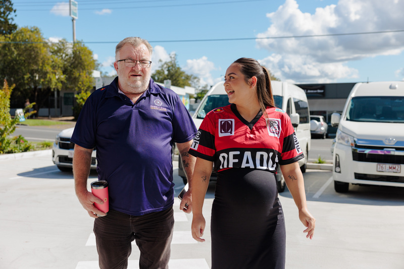 A man and a pregnant woman walk together in a parking lot on a sunny day. The man holds a pink tumbler, and the woman wears a red and black Deadly Choices sports jersey, smiling at him. Several white vans are parked behind them representing the Transport service.