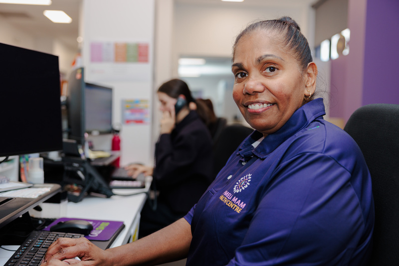 A Pamela Mam Health Centre receptionist wearing a purple uniform with the clinics logo on the front, smiling at the camera.