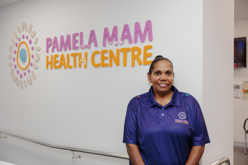 A Pamela Mam Health Centre staff member, wearing the clinics uniform with the logo present on the front of it, standing in front of the clinics sign inside.