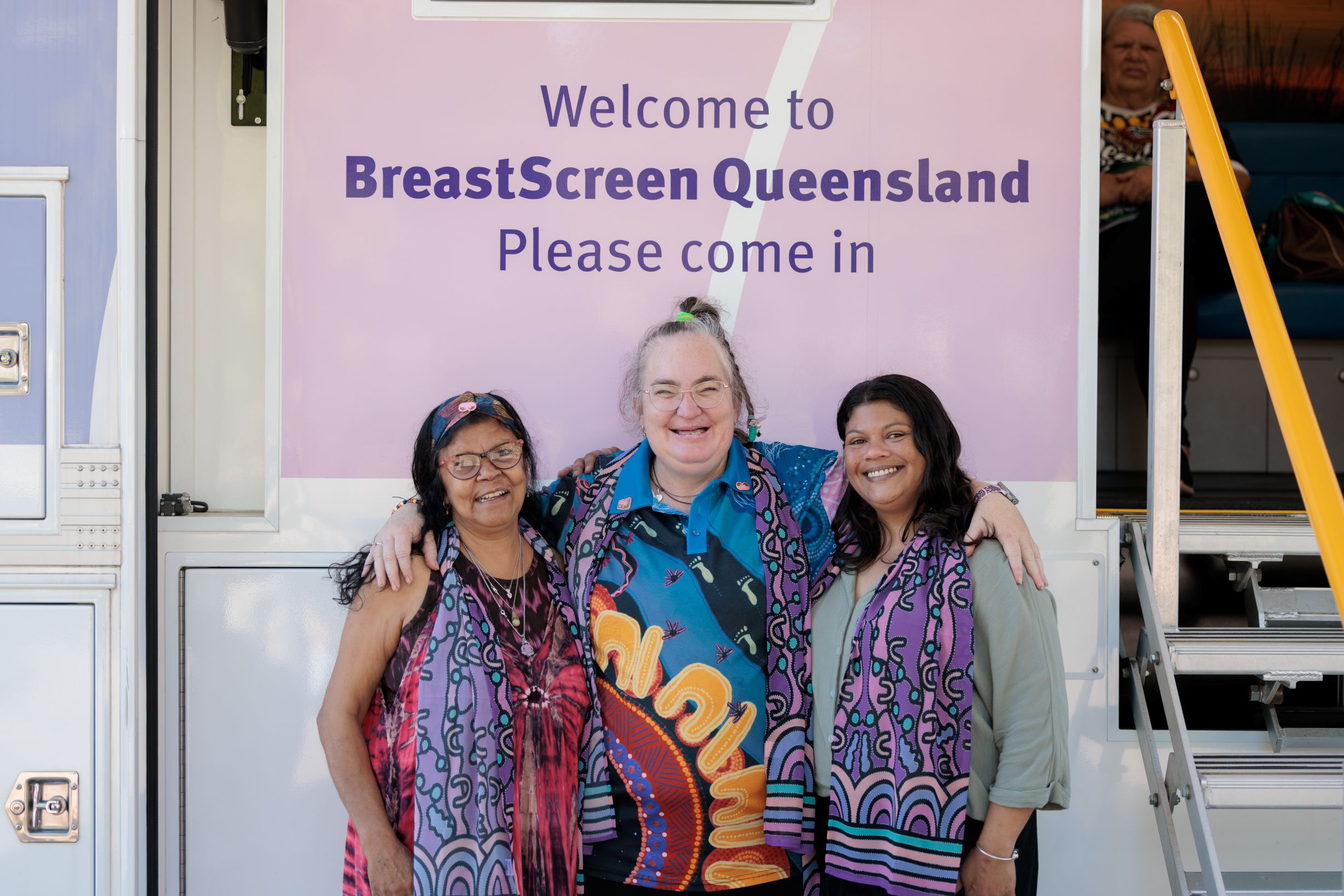 Three smiling women stand together in front of a BreastScreen Queensland sign, wearing colorful clothing with Indigenous patterns to promote breast cancer screening. One woman stands in the middle, arm around each of the others.
