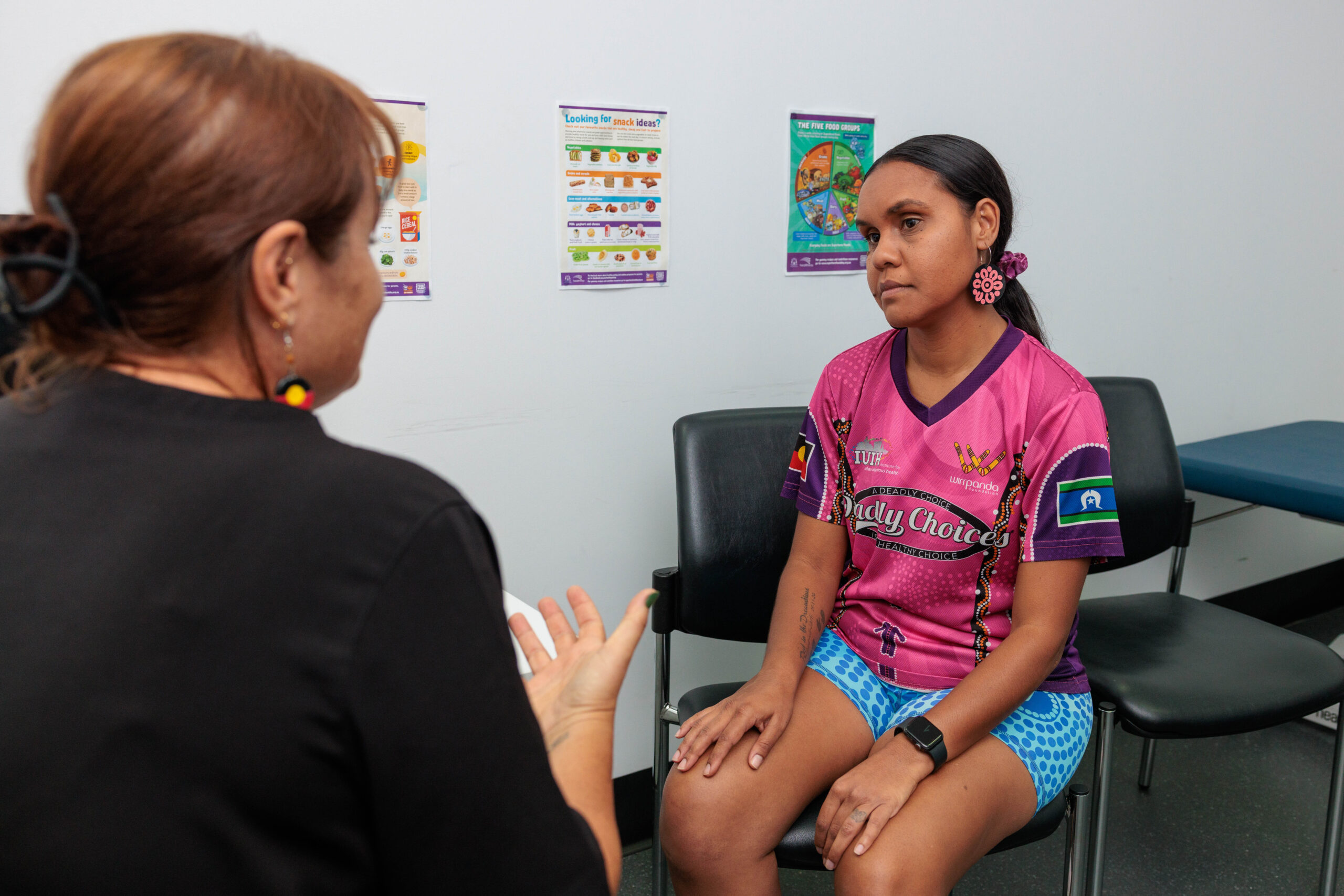 A healthcare professional discusses bowel cancer screening with a woman in a pink shirt sitting on a chair in a clinic room. Health posters are visible on the white wall behind them.